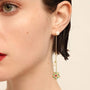 A close-up view of a woman's profile featuring a long, asymmetrical gold earring with a stud pearl at the lobe and a chain extending down to a sunburst pendant encrusted with multicolored gemstones, ending with another pearl. The earring combines classic elegance with a modern, playful design.