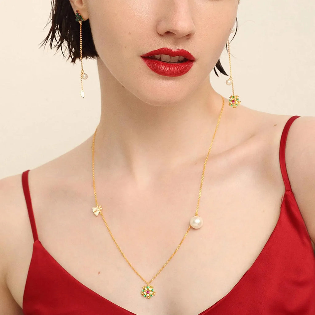 A woman in a red camisole showcases a delicate gold necklace with a small butterfly charm and a larger sunburst pendant adorned with multicolored gemstones, connected by a thin chain with a single pearl. She also wears long, slender gold earrings with a green gem at the stud and a pearl at the end.