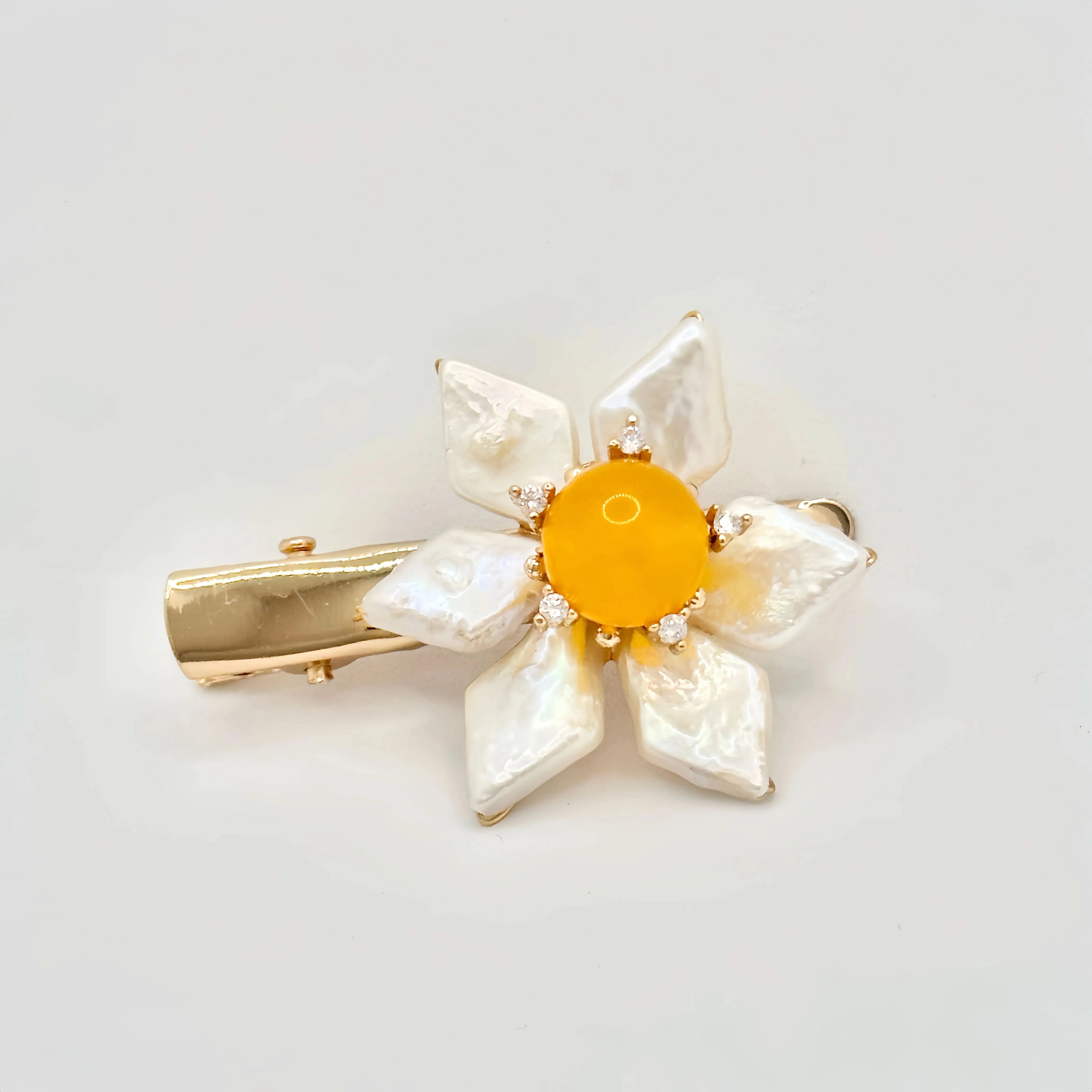 The image features a hair clip with a bright yellow center, surrounded by white baroque pearl petals. Each pearl petal is irregular in shape, adding a natural and organic feel to the design. The center of the flower is accented with small, sparkling stones that complement the gold-tone of the clip's hardware. The overall design resembles a sunny flower, set against a plain background to emphasize its vivid color and lustrous pearls.
