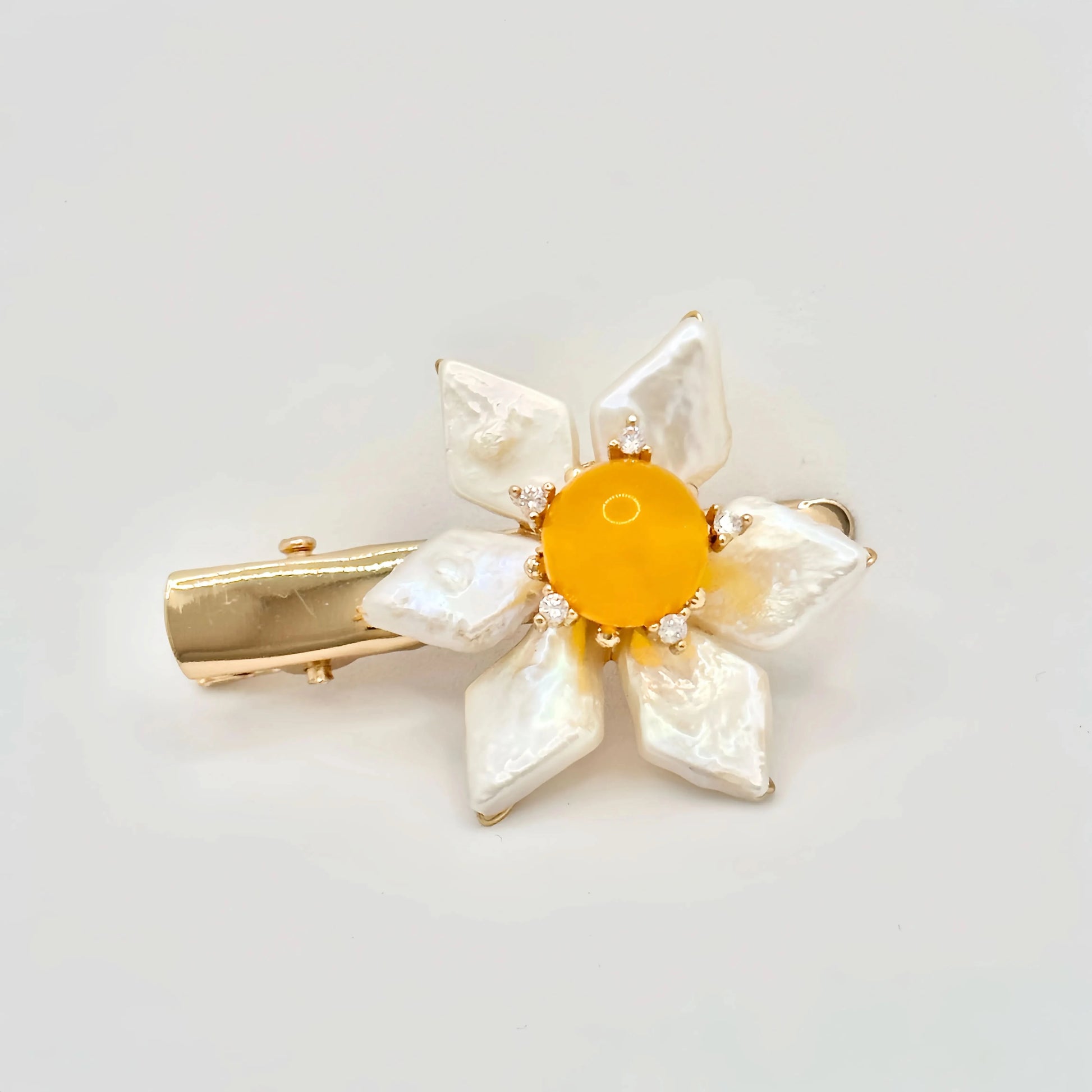 The image features a hair clip with a bright yellow center, surrounded by white baroque pearl petals. Each pearl petal is irregular in shape, adding a natural and organic feel to the design. The center of the flower is accented with small, sparkling stones that complement the gold-tone of the clip's hardware. The overall design resembles a sunny flower, set against a plain background to emphasize its vivid color and lustrous pearls.