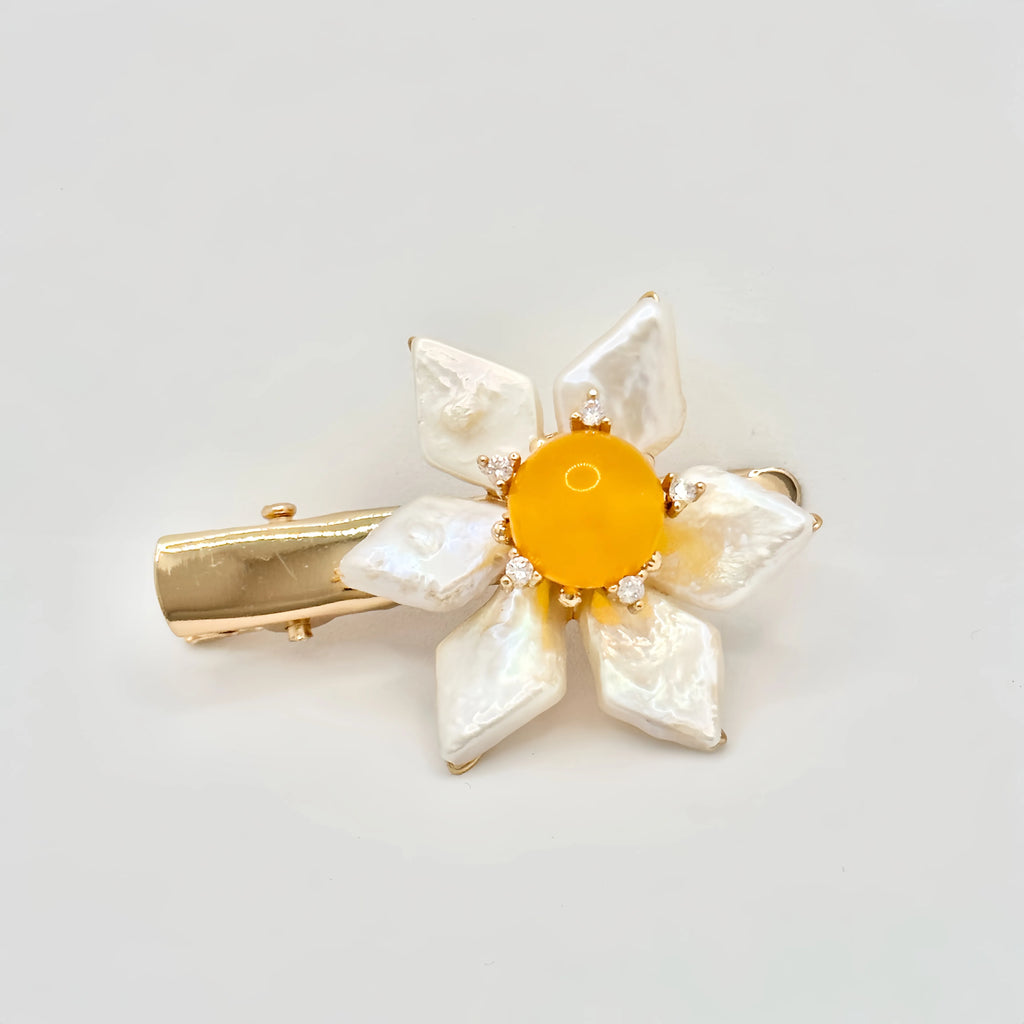 The image features a hair clip with a bright yellow center, surrounded by white baroque pearl petals. Each pearl petal is irregular in shape, adding a natural and organic feel to the design. The center of the flower is accented with small, sparkling stones that complement the gold-tone of the clip's hardware. The overall design resembles a sunny flower, set against a plain background to emphasize its vivid color and lustrous pearls.