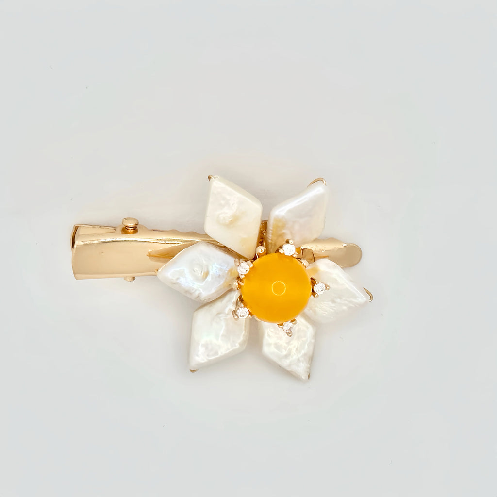 The image features a hair clip with a bright yellow center, surrounded by white baroque pearl petals. Each pearl petal is irregular in shape, adding a natural and organic feel to the design. The center of the flower is accented with small, sparkling stones that complement the gold-tone of the clip's hardware. The overall design resembles a sunny flower, set against a plain background to emphasize its vivid color and lustrous pearls.