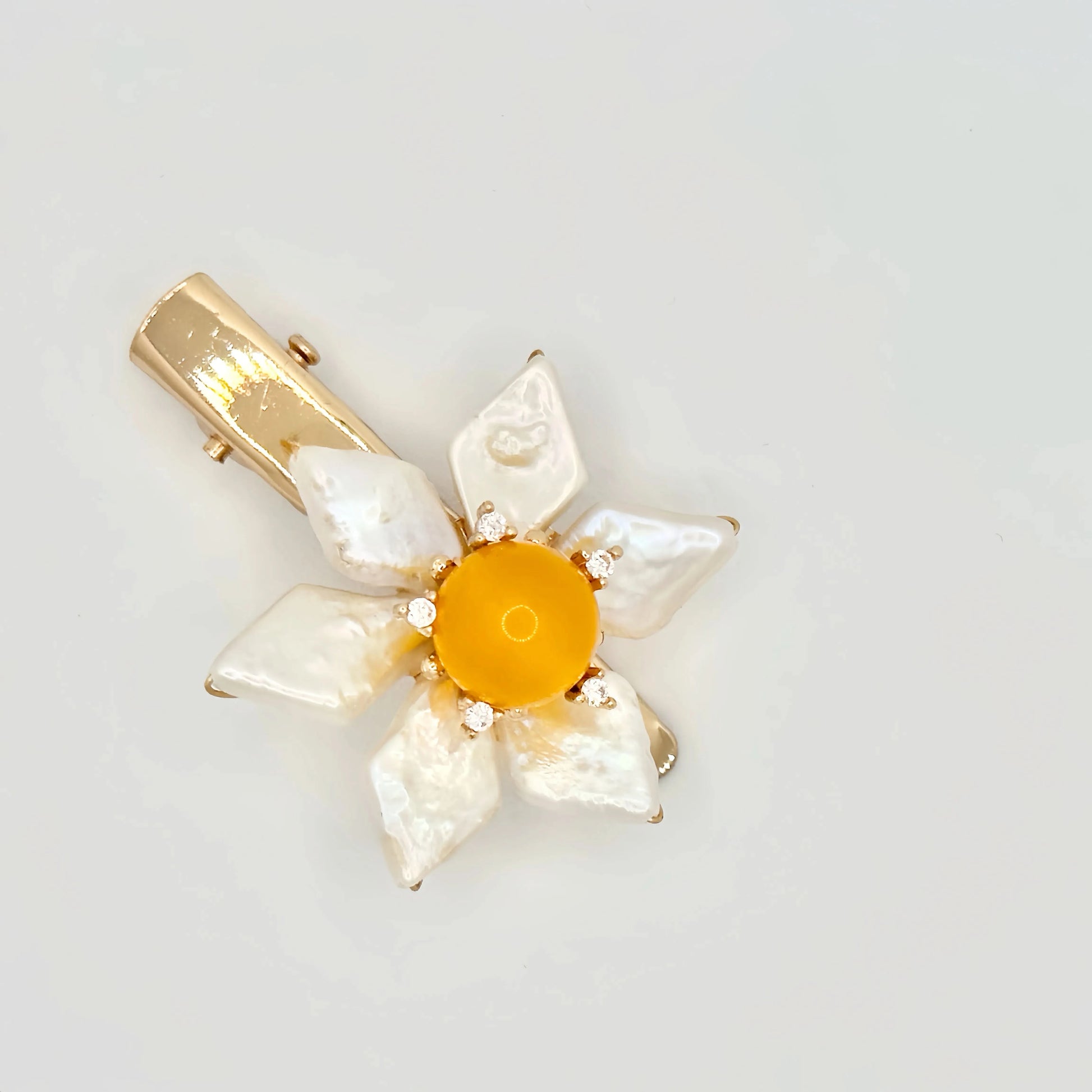The image features a hair clip with a bright yellow center, surrounded by white baroque pearl petals. Each pearl petal is irregular in shape, adding a natural and organic feel to the design. The center of the flower is accented with small, sparkling stones that complement the gold-tone of the clip's hardware. The overall design resembles a sunny flower, set against a plain background to emphasize its vivid color and lustrous pearls.