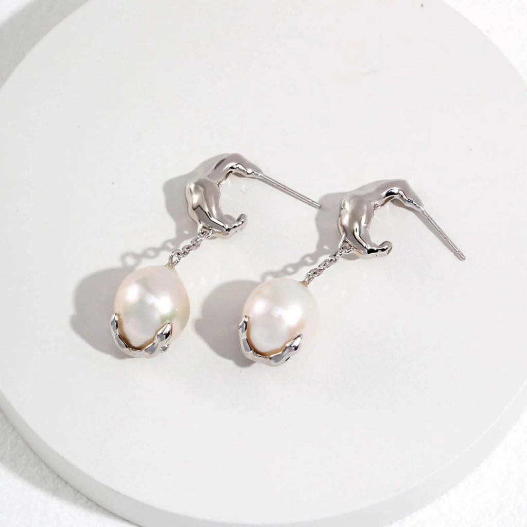 The image displays a pair of gold earrings, each featuring an abstract, sculptural form reminiscent of a human figure bending forward, which serves as the post. A chain links this figure to a large white pearl encased in a gold setting that appears to cradle the pearl gently. The pearls have a soft luster and their organic shape adds a natural elegance to the design. 