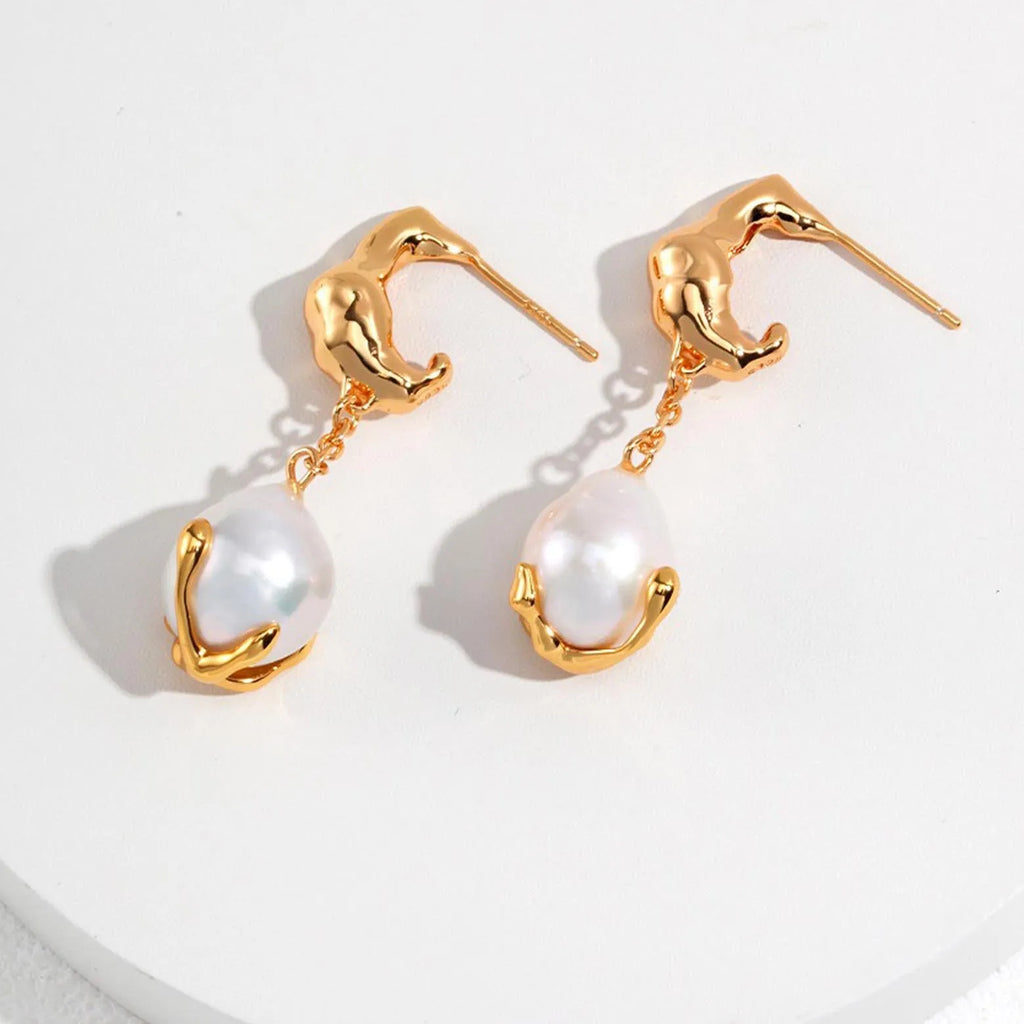 The image displays a pair of gold earrings, each featuring an abstract, sculptural form reminiscent of a human figure bending forward, which serves as the post. A chain links this figure to a large white pearl encased in a gold setting that appears to cradle the pearl gently. The pearls have a soft luster and their organic shape adds a natural elegance to the design. 