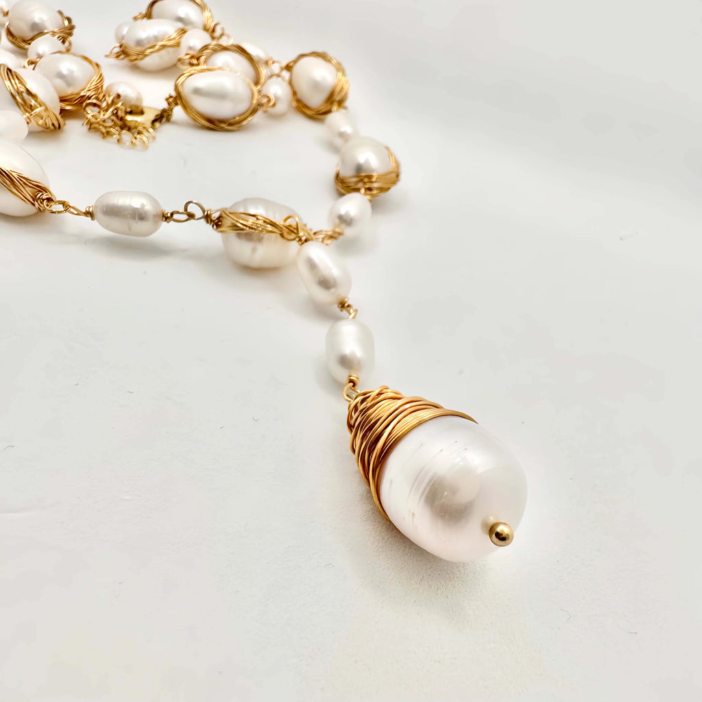 A luxurious pearl jewelry set on a white surface, consisting of a necklace and bracelet. Both pieces feature large, lustrous pearls encased in twisted gold wire, interspersed with smaller pearls linked by gold chains. The necklace is highlighted by a large, baroque pearl pendant, wrapped in a dense coil of gold wire, adding a striking visual element to the set.