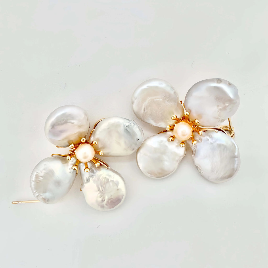 A gold brooch with a floral design, featuring large, baroque pearls as petals, a smaller central pearl, and golden balls accentuating the center and edges. The lustrous pearls have a natural, irregular shape, which adds to the unique and organic feel of the piece, set against a white background.