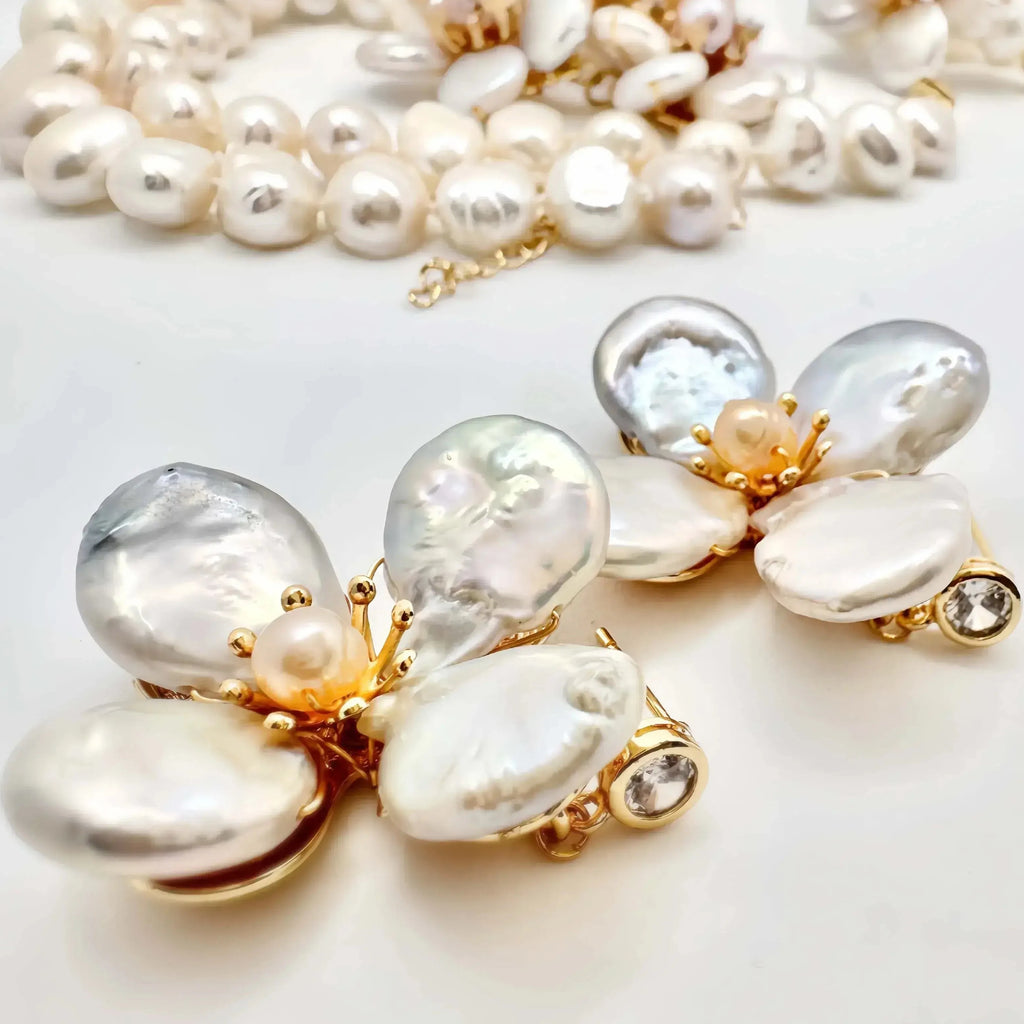 An exquisite pearl jewelry set displayed on a white background, featuring a necklace and a statement brooch. The necklace is made up of multiple strands of creamy white pearls of various sizes. The brooch is designed as a flower, with irregularly shaped pearl petals and gold accents, highlighted by small sparkling stones at the base of each petal.
