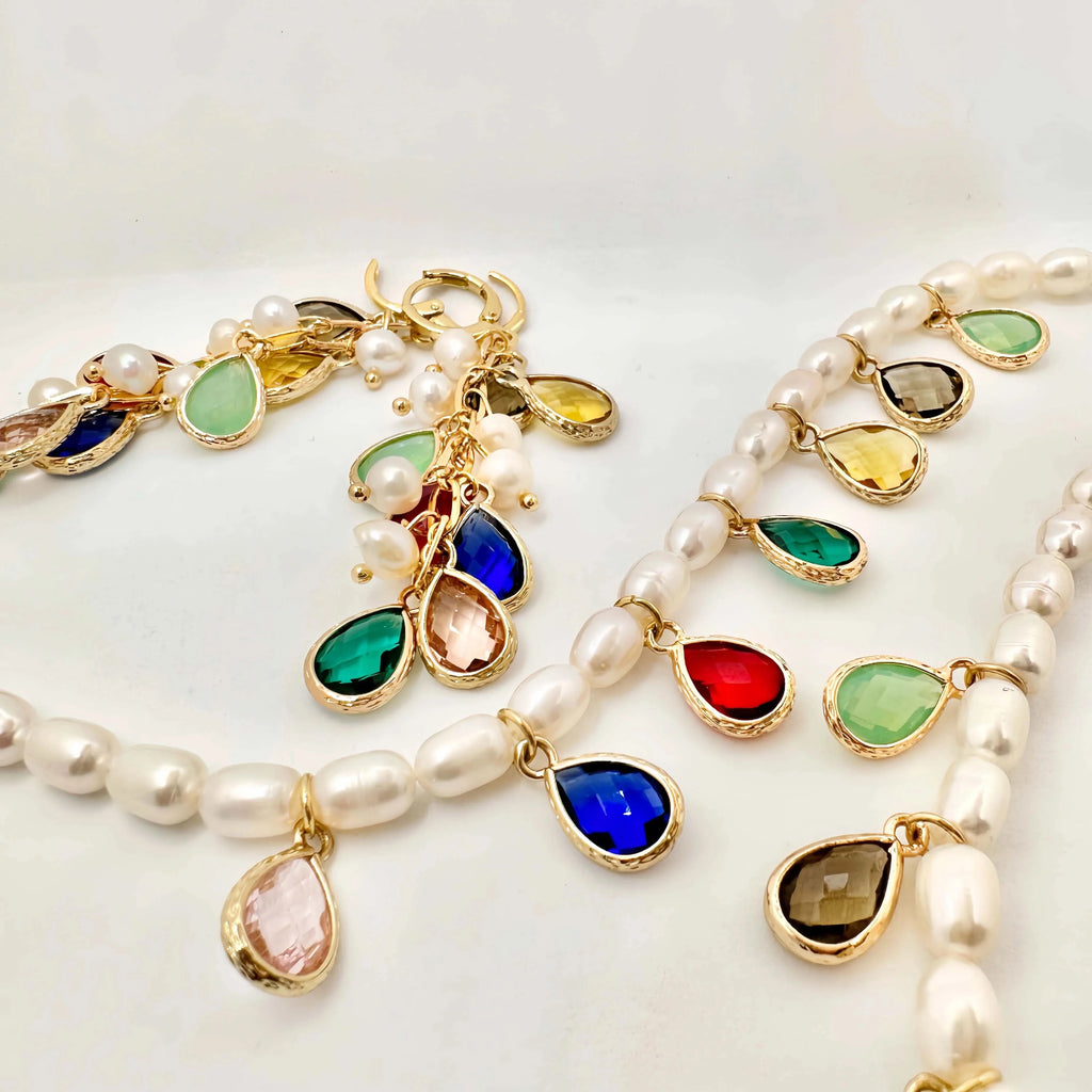 A luxurious pearl necklace and bracelet set displayed on a white background. The necklace is a string of round white pearls, interspersed with gold links and teardrop-shaped colored gemstones in bezel settings. The bracelet mirrors the design with similar colored gems and smaller pearls, both creating a harmonious and opulent look.