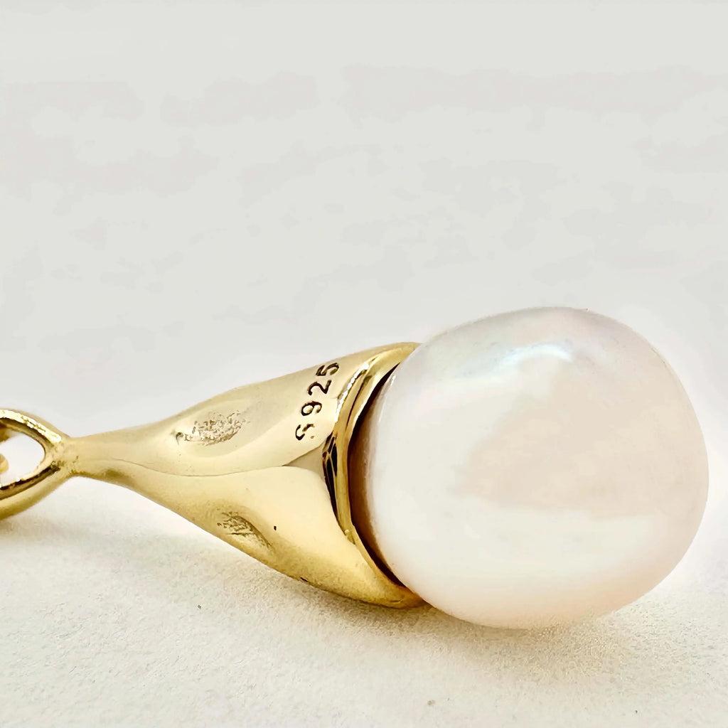 The image displays a close-up of a gold pendant earring with a large, round, white pearl. The pearl is set in a gold cap which connects to a delicate gold chain. The gold has a polished finish and is marked with "925," indicating that the metal is sterling silver that has been gold-plated. The pearl exhibits a soft luster and a smooth surface, reflecting the light in a gentle glow. The background is a soft, neutral color, which complements the warmth of the gold and the serene beauty of the pearl.