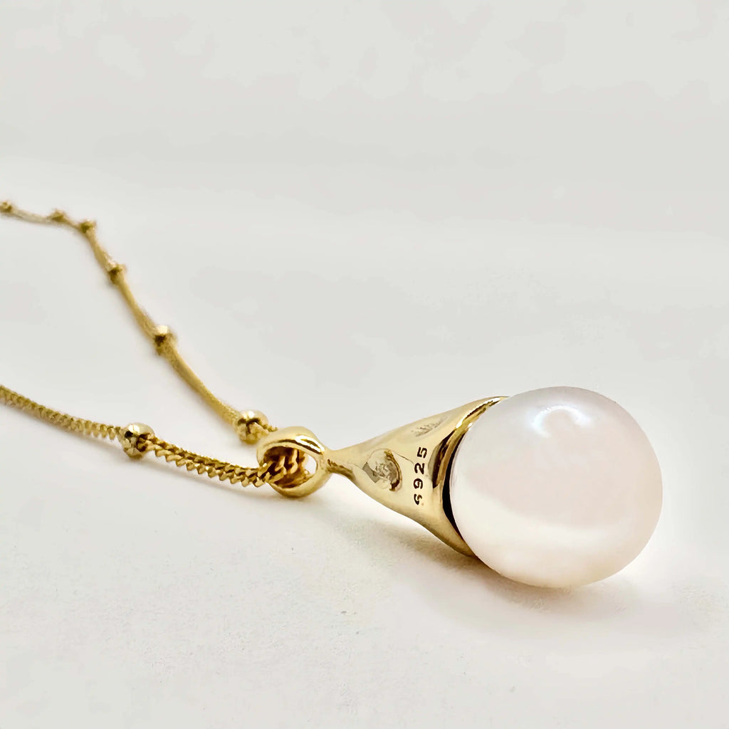 The image displays a close-up of a gold pendant earring with a large, round, white pearl. The pearl is set in a gold cap which connects to a delicate gold chain. The gold has a polished finish and is marked with "925," indicating that the metal is sterling silver that has been gold-plated. The pearl exhibits a soft luster and a smooth surface, reflecting the light in a gentle glow. The background is a soft, neutral color, which complements the warmth of the gold and the serene beauty of the pearl.