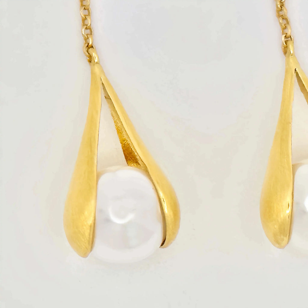 The image depicts a pair of elegant drop earrings featuring large, white baroque pearls. Each pearl is held within a gold setting that gently hugs the pearl's irregular shape, giving the impression of being lovingly cradled. The gold has a brushed, matte finish that contrasts with the smooth, glossy surface of the pearls.