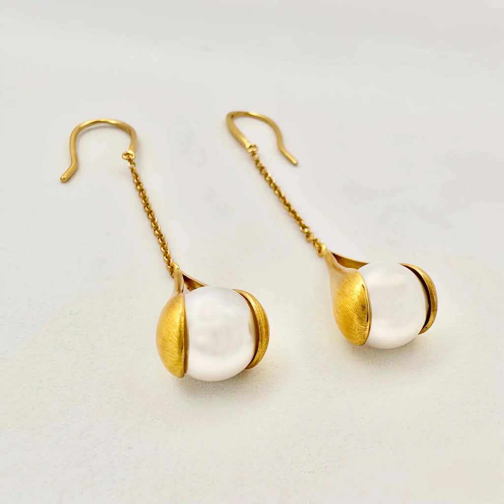 The image depicts a pair of elegant drop earrings featuring large, white baroque pearls. Each pearl is held within a gold setting that gently hugs the pearl's irregular shape, giving the impression of being lovingly cradled. The gold has a brushed, matte finish that contrasts with the smooth, glossy surface of the pearls.