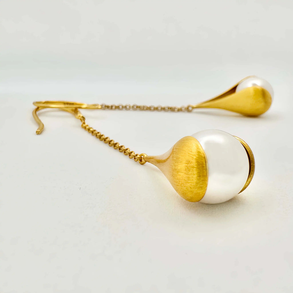 The image depicts a pair of elegant drop earrings featuring large, white baroque pearls. Each pearl is held within a gold setting that gently hugs the pearl's irregular shape, giving the impression of being lovingly cradled. The gold has a brushed, matte finish that contrasts with the smooth, glossy surface of the pearls.