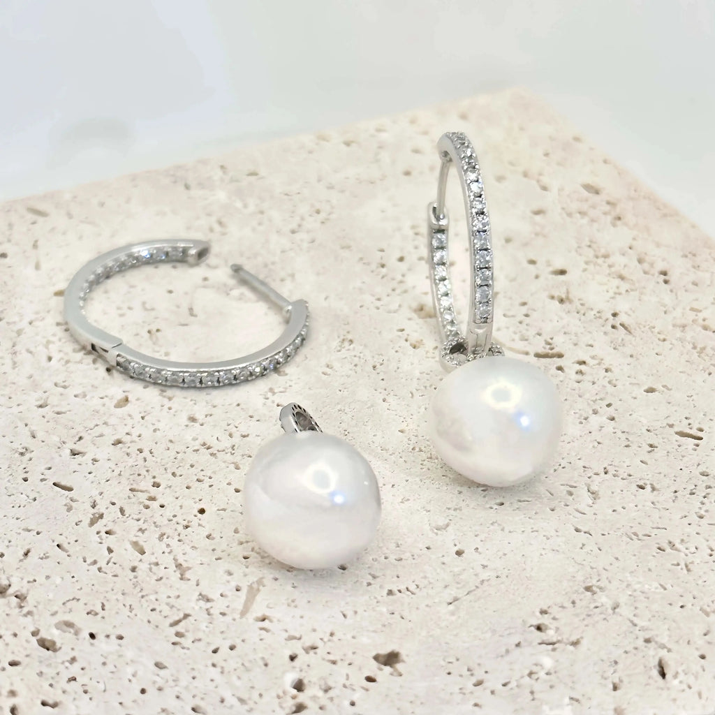 This image shows a pair of silver hoop earrings, each lined with small, clear crystals that provide a sparkling accent. Below the hoops, there are two large, white pearls with a notable sheen, reflecting light and showing hints of iridescence. The pearls are detached from the hoops, suggesting they may be interchangeable or part of a set. The earrings and pearls rest against a textured, beige surface that resembles sandstone, allowing the jewelry's polished finish to stand out.