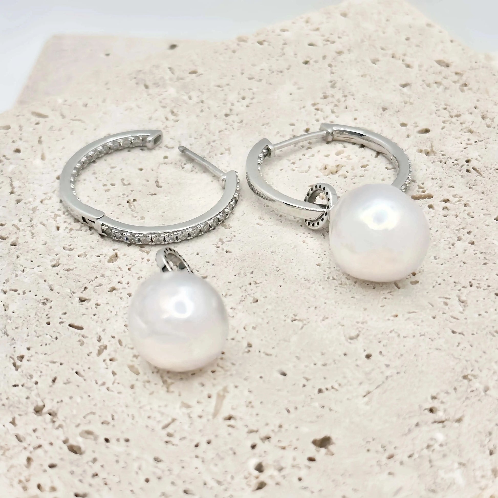 This image shows a pair of silver hoop earrings, each lined with small, clear crystals that provide a sparkling accent. Below the hoops, there are two large, white pearls with a notable sheen, reflecting light and showing hints of iridescence. The pearls are detached from the hoops, suggesting they may be interchangeable or part of a set. The earrings and pearls rest against a textured, beige surface that resembles sandstone, allowing the jewelry's polished finish to stand out.