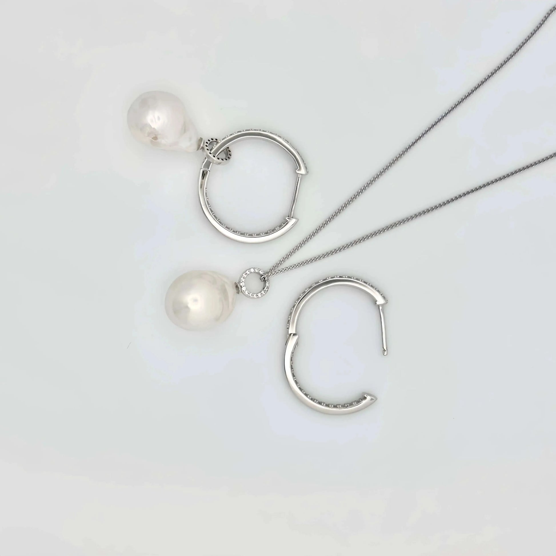 This image shows a pair of silver hoop earrings, each lined with small, clear crystals that provide a sparkling accent. Below the hoops, there are two large, white pearls with a notable sheen, reflecting light and showing hints of iridescence. The pearls are detached from the hoops, suggesting they may be interchangeable or part of a set. The earrings and pearls rest against a textured, beige surface that resembles sandstone, allowing the jewelry's polished finish to stand out.