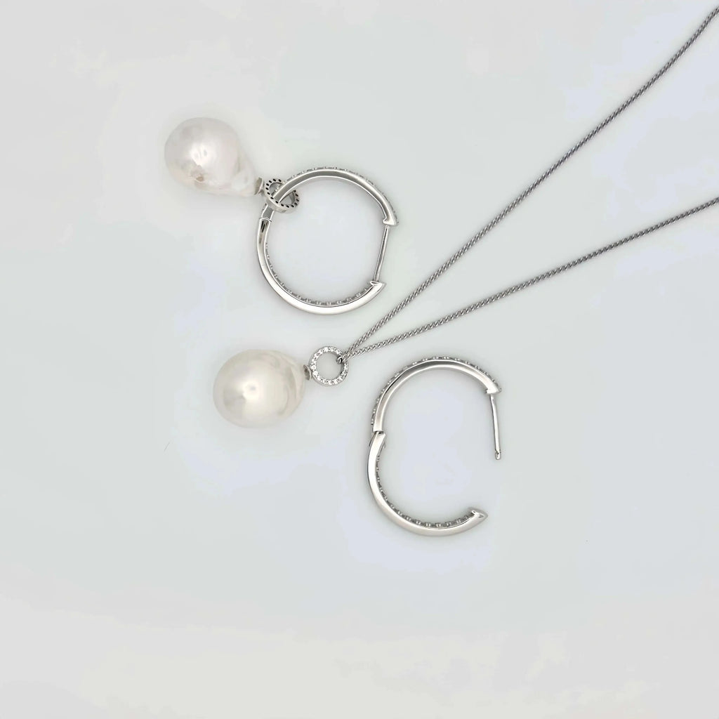 This image shows a pair of silver hoop earrings, each lined with small, clear crystals that provide a sparkling accent. Below the hoops, there are two large, white pearls with a notable sheen, reflecting light and showing hints of iridescence. The pearls are detached from the hoops, suggesting they may be interchangeable or part of a set. The earrings and pearls rest against a textured, beige surface that resembles sandstone, allowing the jewelry's polished finish to stand out.