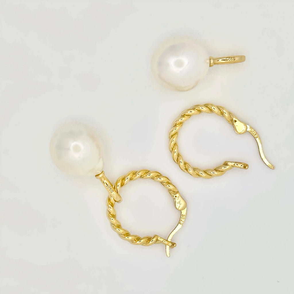 This image features a pair of gold earrings, each adorned with a large, lustrous white pearl. The pearls exhibit a smooth, round shape with a glossy finish, appearing well-matched in size. The metallic components of the earrings are gold in color, suggesting high-quality metal, possibly gold, crafted in a simple yet elegant twisted chain design. The background is a muted, light color, accentuating the jewelry's color and sheen.