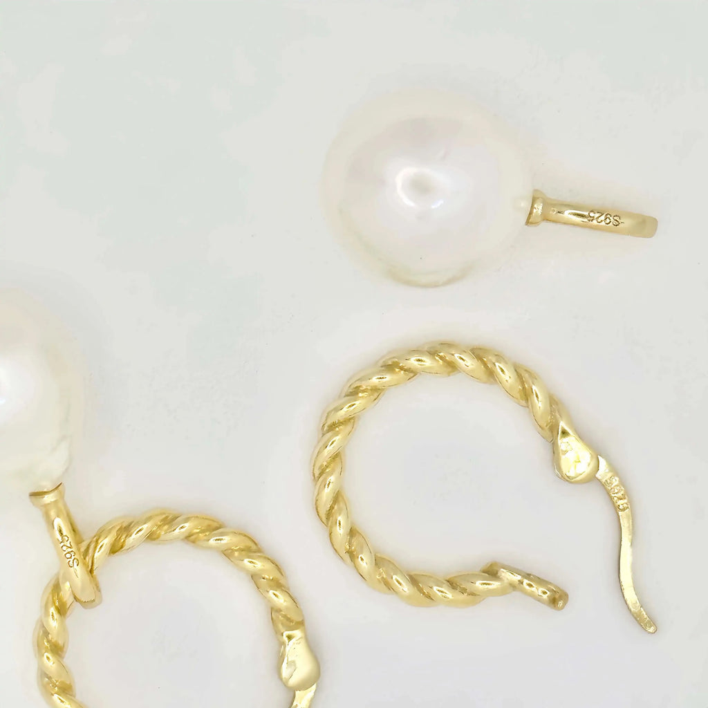 This image features a pair of gold earrings, each adorned with a large, lustrous white pearl. The pearls exhibit a smooth, round shape with a glossy finish, appearing well-matched in size. The metallic components of the earrings are gold in color, suggesting high-quality metal, possibly gold, crafted in a simple yet elegant twisted chain design. The background is a muted, light color, accentuating the jewelry's color and sheen.