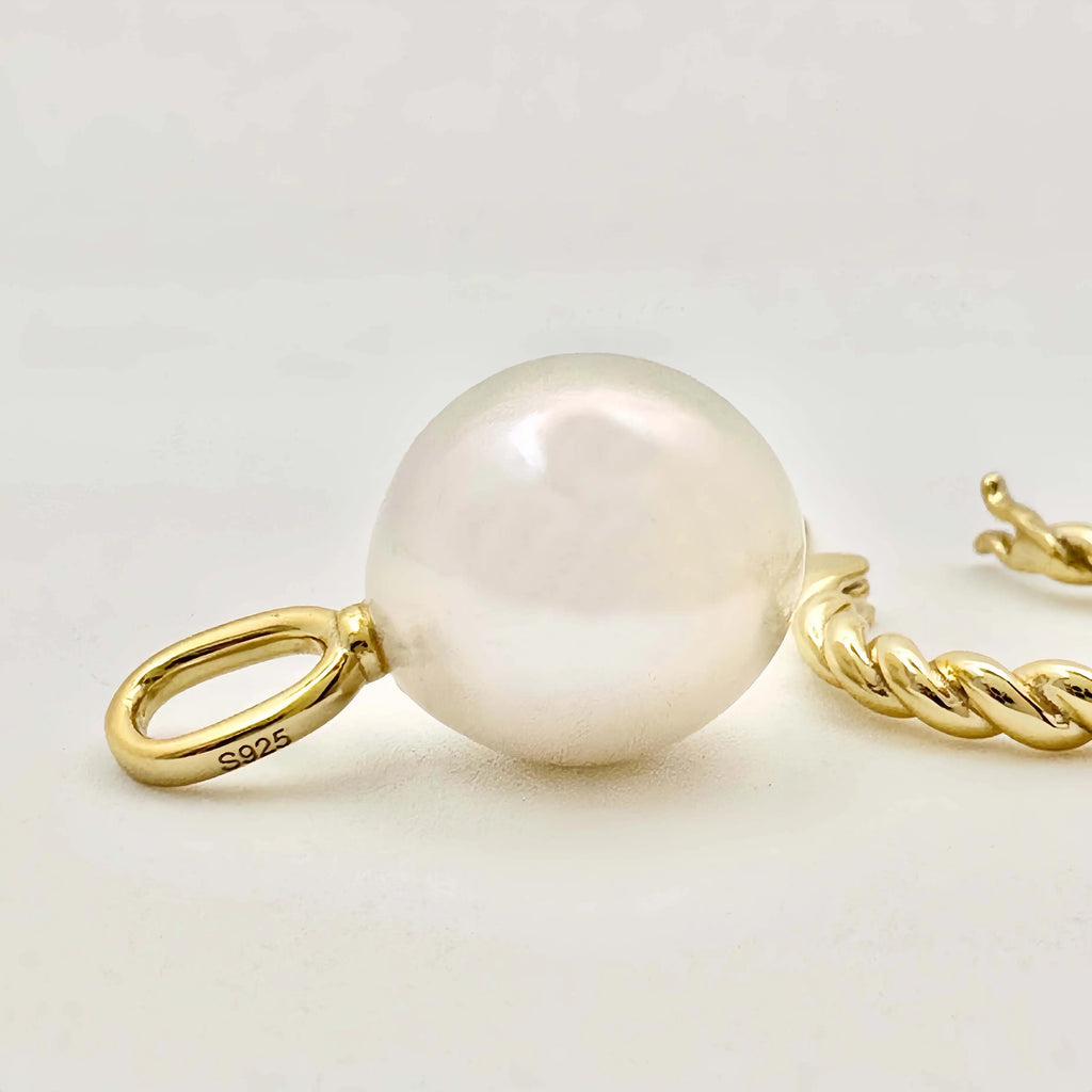 This image features a pair of gold earrings, each adorned with a large, lustrous white pearl. The pearls exhibit a smooth, round shape with a glossy finish, appearing well-matched in size. The metallic components of the earrings are gold in color, suggesting high-quality metal, possibly gold, crafted in a simple yet elegant twisted chain design. The background is a muted, light color, accentuating the jewelry's color and sheen.