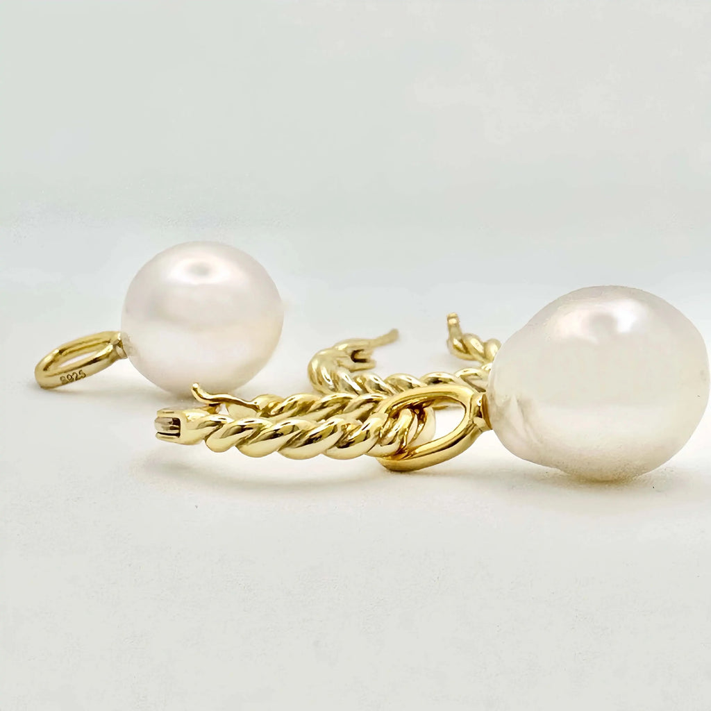 This image features a pair of gold earrings, each adorned with a large, lustrous white pearl. The pearls exhibit a smooth, round shape with a glossy finish, appearing well-matched in size. The metallic components of the earrings are gold in color, suggesting high-quality metal, possibly gold, crafted in a simple yet elegant twisted chain design. The background is a muted, light color, accentuating the jewelry's color and sheen.