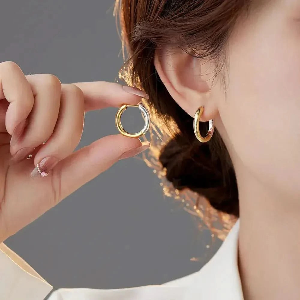 A woman is shown from the side, gently holding a small gold hoop earring with her fingers, preparing to adorn her ear with its mate, which is already in place. The earrings are simple yet elegant, reflecting the light and complementing her sophisticated, minimalistic style.