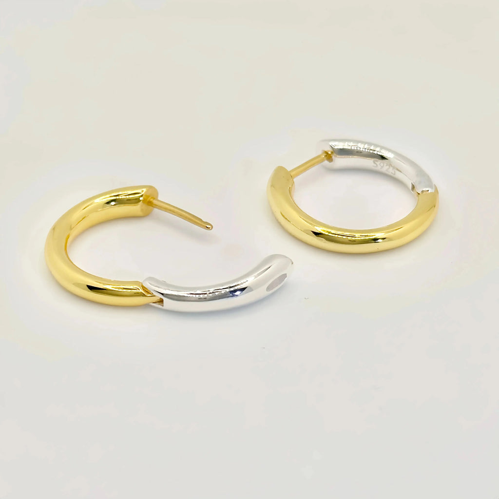 A woman is shown from the side, gently holding a small gold hoop earring with her fingers, preparing to adorn her ear with its mate, which is already in place. The earrings are simple yet elegant, reflecting the light and complementing her sophisticated, minimalistic style.