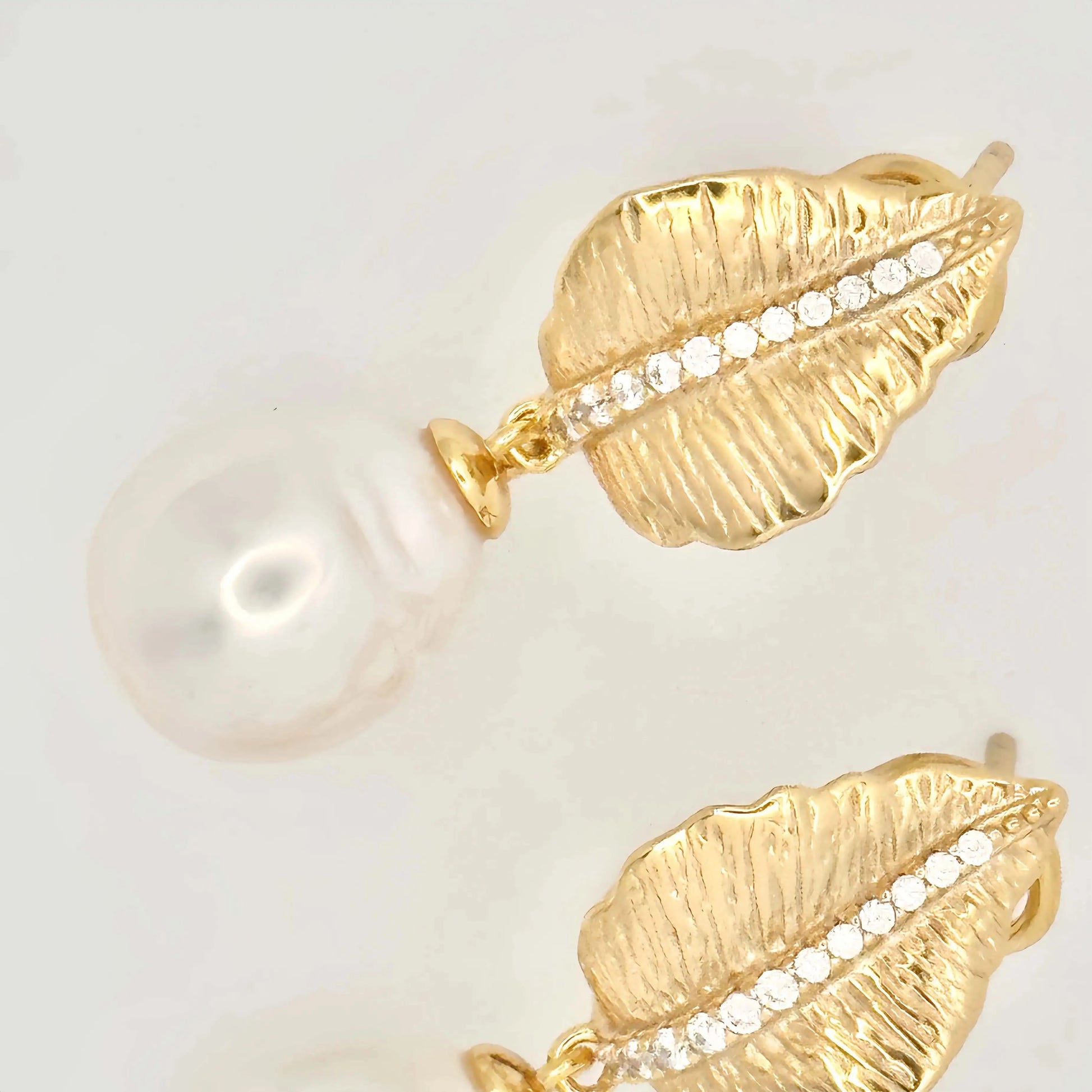 A pair of sophisticated earrings with a nature-inspired design, featuring large white pearls. Each pearl is mounted on a gold setting that resembles a textured leaf or petal, accented with a line of small diamonds that add sparkle to the organic motif, all set against a neutral background.