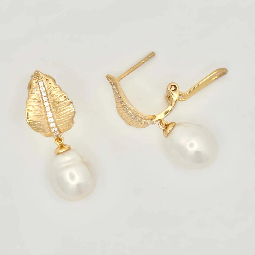 A pair of sophisticated earrings with a nature-inspired design, featuring large white pearls. Each pearl is mounted on a gold setting that resembles a textured leaf or petal, accented with a line of small diamonds that add sparkle to the organic motif, all set against a neutral background.