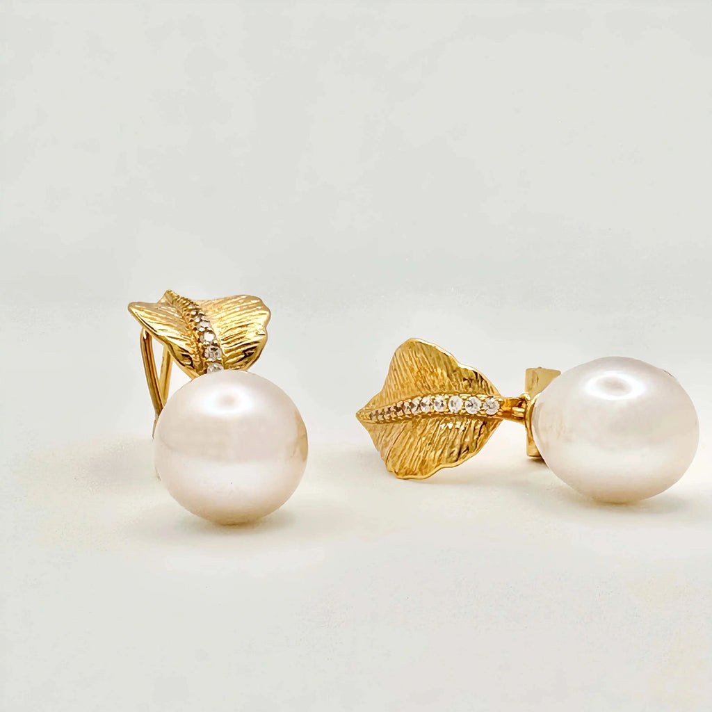 A pair of sophisticated earrings with a nature-inspired design, featuring large white pearls. Each pearl is mounted on a gold setting that resembles a textured leaf or petal, accented with a line of small diamonds that add sparkle to the organic motif, all set against a neutral background.