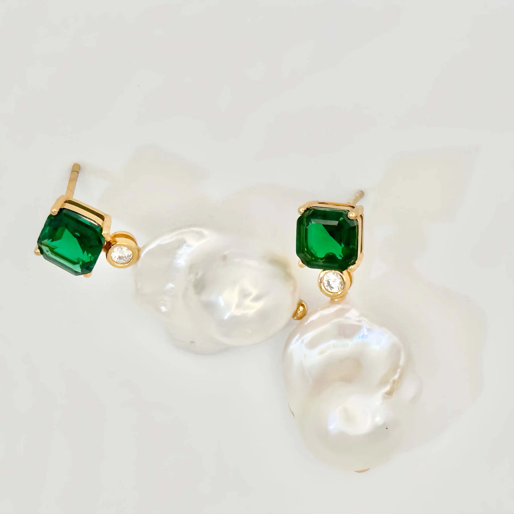 A pair of elegant earrings presented on a white background. Each earring features a large square-cut green gemstone set in gold, with a small round diamond below it, leading to an irregularly shaped baroque pearl that adds a touch of organic sophistication to the design.