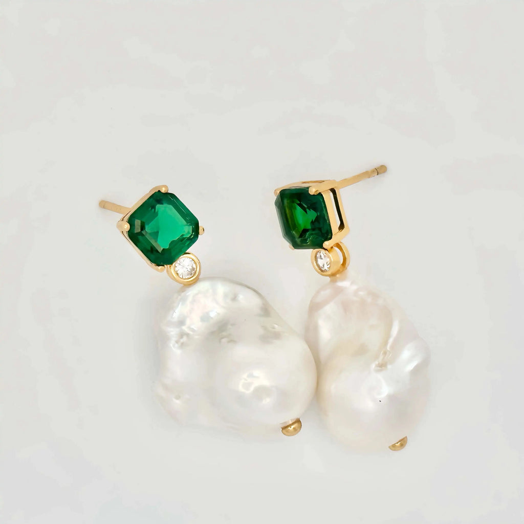 A pair of elegant earrings presented on a white background. Each earring features a large square-cut green gemstone set in gold, with a small round diamond below it, leading to an irregularly shaped baroque pearl that adds a touch of organic sophistication to the design.