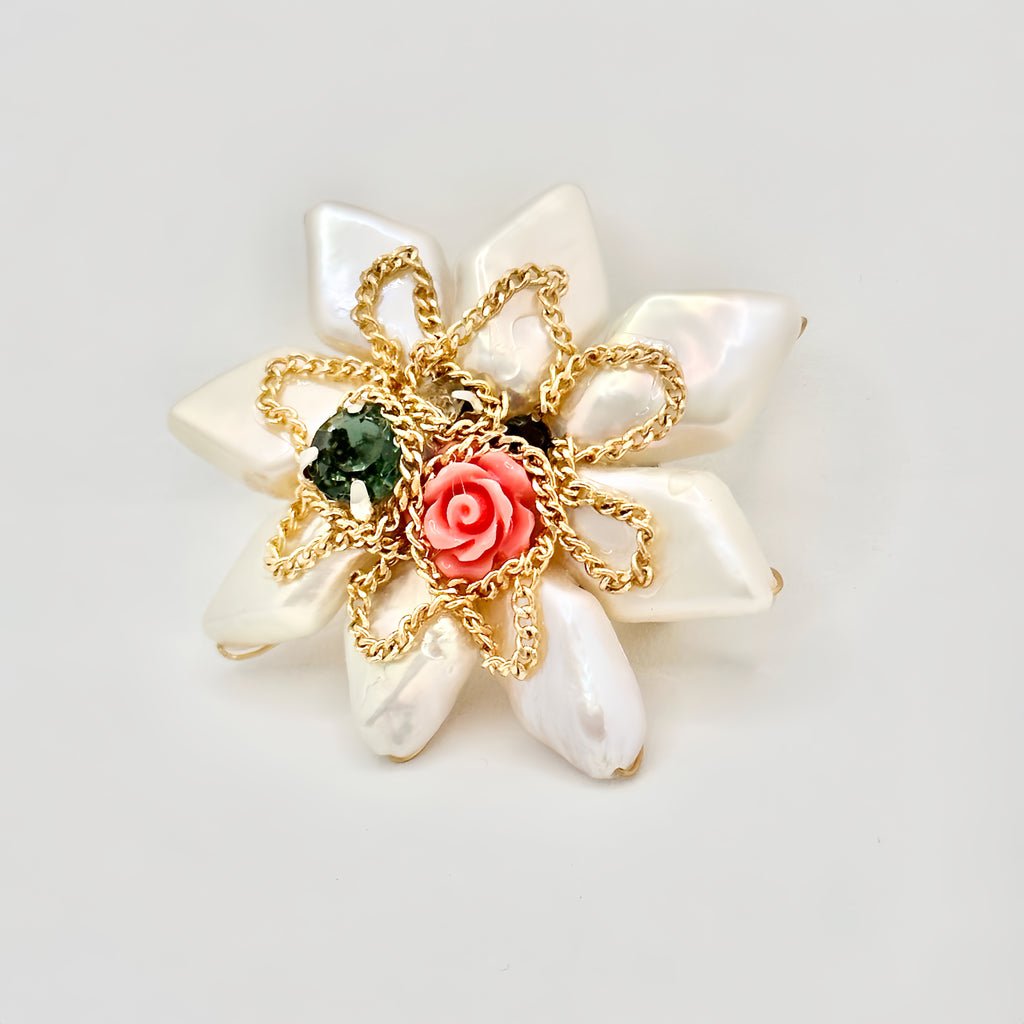 The image depicts a gold-toned hair clip with a large, star-shaped design crafted from iridescent baroque pearls. At the center is a detailed pink rose, encircled by a gold chain loop. Adjacent to the rose is a single, dark green gemstone, adding a contrasting pop of color. The overall design suggests a blend of natural elegance and ornate detailing, presented against a clear, neutral background.