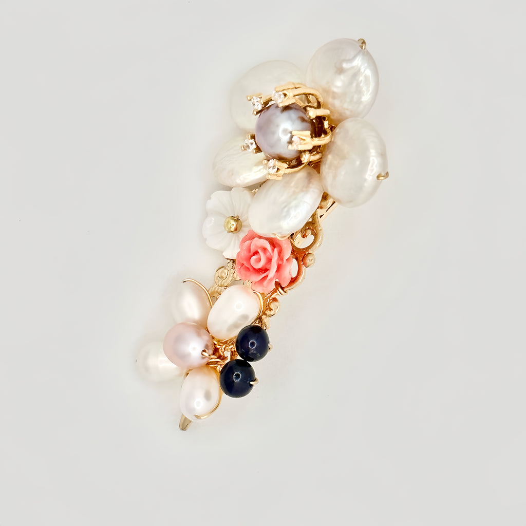 The image presents a gold-toned hair clip adorned with a variety of pearls and a single pink rose. The pearls are in different shades and sizes, including large white baroque pearls, smaller pink and dark blue pearls, and one prominent pearl with a ring of gold and tiny crystal accents. The pink rose adds a soft, romantic touch to the clip, while the arrangement of pearls gives it a luxurious and eclectic charm. The clip is set against a plain background that highlights its delicate and ornate composition.