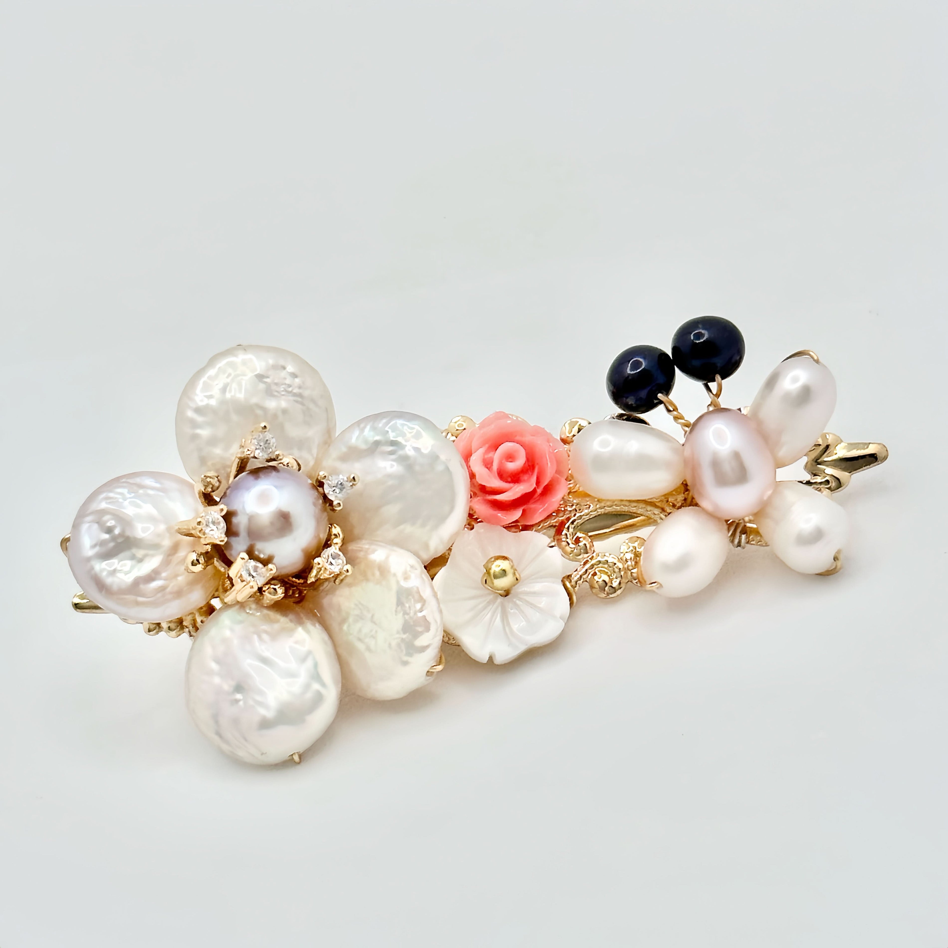 The image presents a gold-toned hair clip adorned with a variety of pearls and a single pink rose. The pearls are in different shades and sizes, including large white baroque pearls, smaller pink and dark blue pearls, and one prominent pearl with a ring of gold and tiny crystal accents. The pink rose adds a soft, romantic touch to the clip, while the arrangement of pearls gives it a luxurious and eclectic charm. The clip is set against a plain background that highlights its delicate and ornate composition.