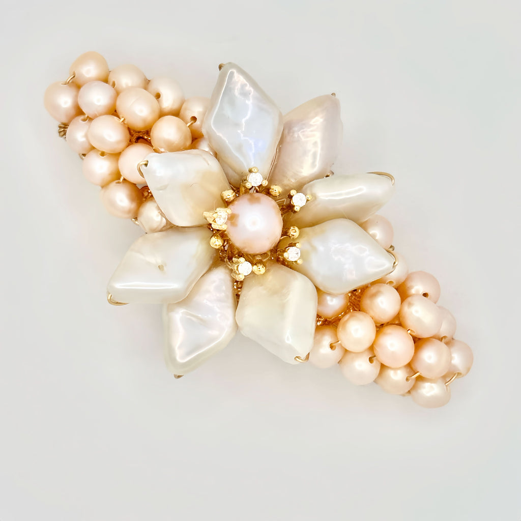 The image shows a golden hair clip adorned with a prominent flower made from large, iridescent baroque pearls. At its center is a smaller, round pearl encircled by tiny gold accents with sparkling elements. Surrounding the flower is a cluster of smaller, pastel pink pearls, giving the appearance of a full bloom. The piece is set against a soft, neutral background that enhances the warm, subtle hues of the pearls.