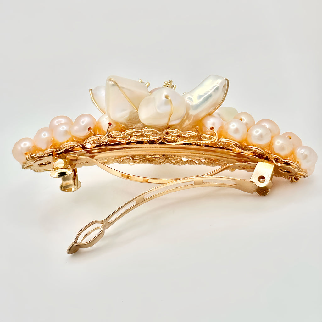 The image shows a golden hair clip adorned with a prominent flower made from large, iridescent baroque pearls. At its center is a smaller, round pearl encircled by tiny gold accents with sparkling elements. Surrounding the flower is a cluster of smaller, pastel pink pearls, giving the appearance of a full bloom. The piece is set against a soft, neutral background that enhances the warm, subtle hues of the pearls.