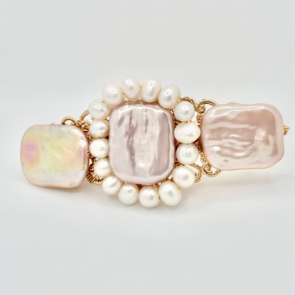 The image shows a gold-toned hair clip adorned with a central rectangular pink pearl, flanked by two irregular-shaped pearls with a lustrous sheen reflecting pastel hues. Surrounding the central pearls is a ring of smaller, round white pearls, set against the intricate gold filigree detailing of the clip. The piece exudes a blend of classic elegance and modern charm, set against a neutral background that highlights its colors and textures.