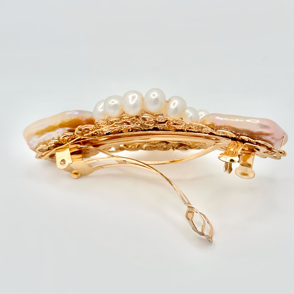 The image shows a gold-toned hair clip adorned with a central rectangular pink pearl, flanked by two irregular-shaped pearls with a lustrous sheen reflecting pastel hues. Surrounding the central pearls is a ring of smaller, round white pearls, set against the intricate gold filigree detailing of the clip. The piece exudes a blend of classic elegance and modern charm, set against a neutral background that highlights its colors and textures.