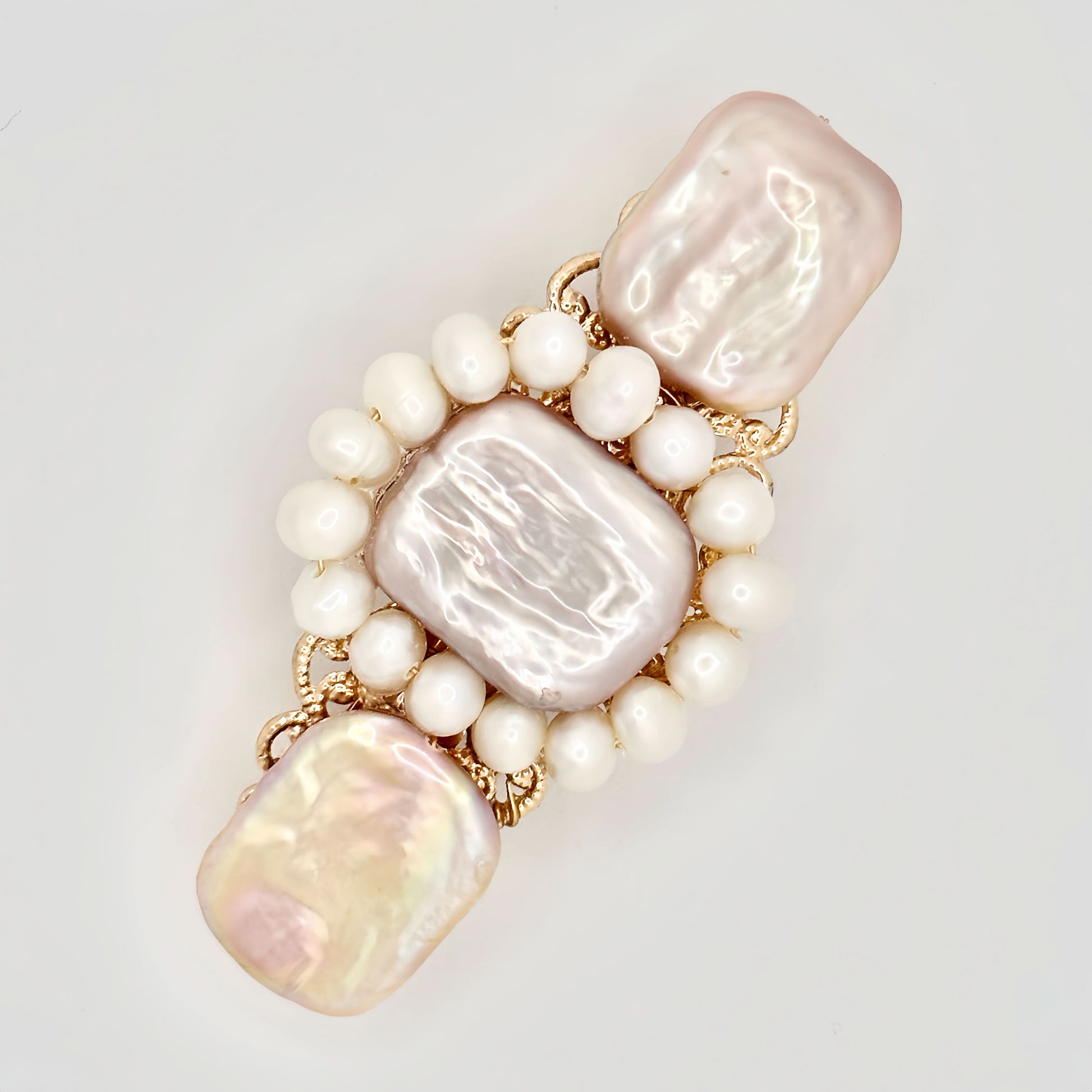 The image shows a gold-toned hair clip adorned with a central rectangular pink pearl, flanked by two irregular-shaped pearls with a lustrous sheen reflecting pastel hues. Surrounding the central pearls is a ring of smaller, round white pearls, set against the intricate gold filigree detailing of the clip. The piece exudes a blend of classic elegance and modern charm, set against a neutral background that highlights its colors and textures.