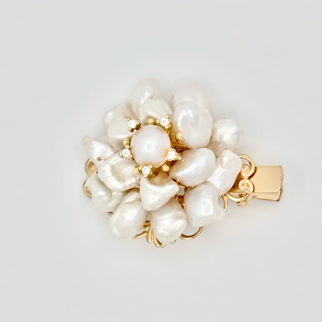 The image showcases a luxurious gold hair clip with a floral motif, featuring a central round pearl surrounded by an array of baroque pearls forming petals. Each pearl petal is edged with tiny gold accents, adding a hint of sparkle. The overall design captures the essence of an opulent blossom, with the golden tones warmly complementing the creamy luster of the pearls. The clip's background is a clean, solid color that accentuates the jewelry's elegant details.