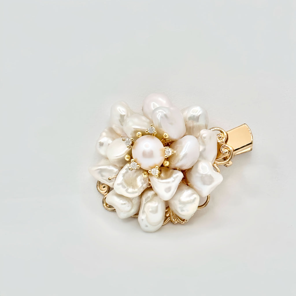 The image showcases a luxurious gold hair clip with a floral motif, featuring a central round pearl surrounded by an array of baroque pearls forming petals. Each pearl petal is edged with tiny gold accents, adding a hint of sparkle. The overall design captures the essence of an opulent blossom, with the golden tones warmly complementing the creamy luster of the pearls. The clip's background is a clean, solid color that accentuates the jewelry's elegant details.