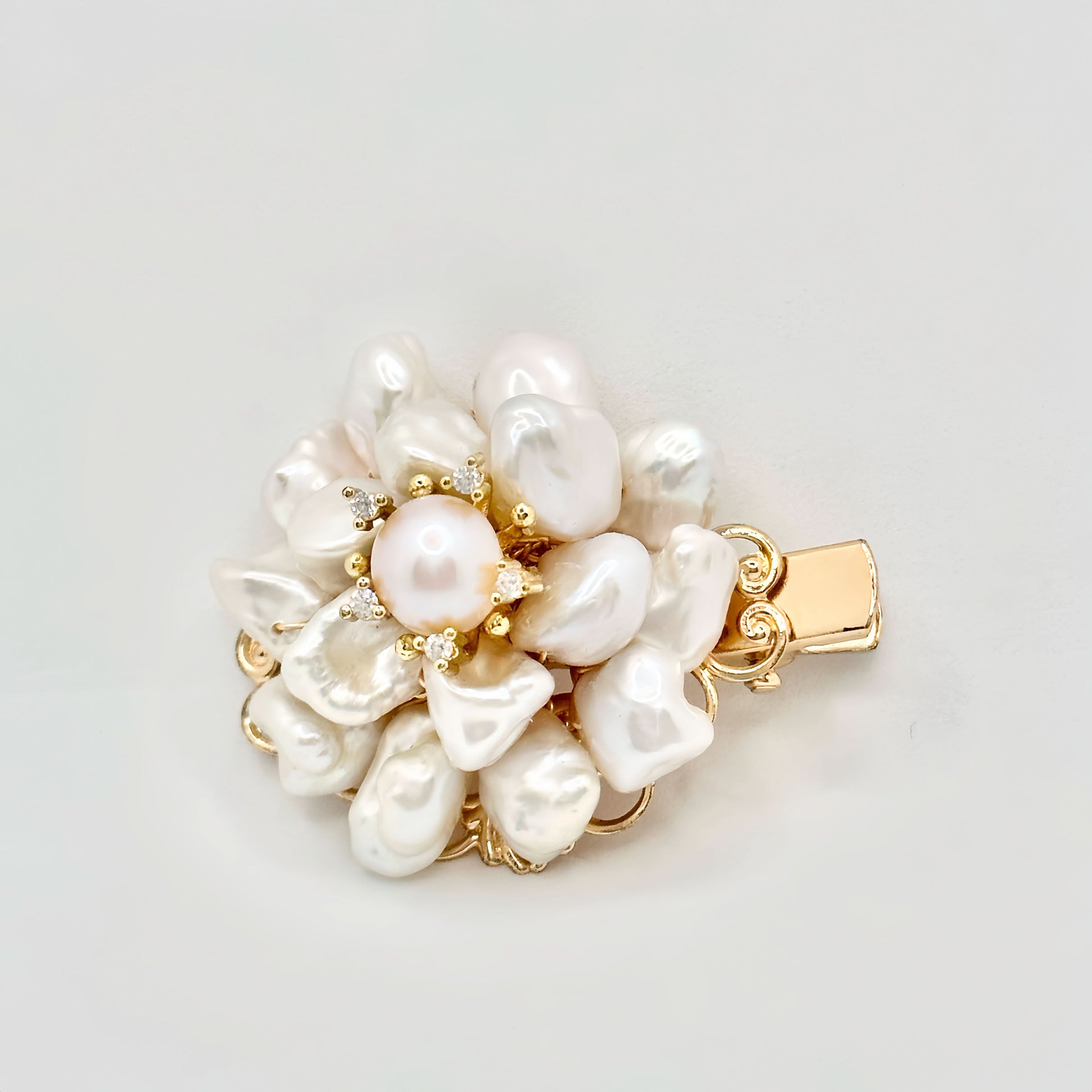 The image showcases a luxurious gold hair clip with a floral motif, featuring a central round pearl surrounded by an array of baroque pearls forming petals. Each pearl petal is edged with tiny gold accents, adding a hint of sparkle. The overall design captures the essence of an opulent blossom, with the golden tones warmly complementing the creamy luster of the pearls. The clip's background is a clean, solid color that accentuates the jewelry's elegant details.