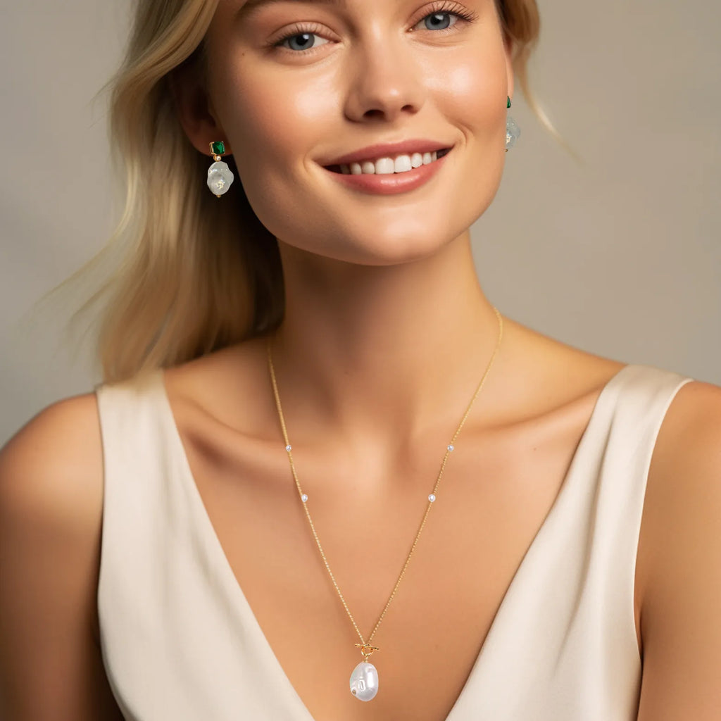 A portrait of a smiling blonde woman with a subtle makeup look, wearing a delicate gold necklace with small white pearls spaced along its length and a larger pearl pendant. She also wears matching earrings, each with a green gemstone stud and a similar large pearl drop, complementing her elegant off-white outfit.