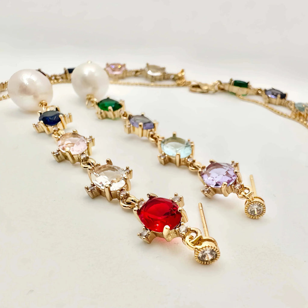 A gold bracelet adorned with a series of colorful gemstones in various cuts and settings, including blue, green, pink, and a prominent red central stone. The bracelet also features two large pearls and smaller round cut gems, culminating in a charm-like design with a dangling round-cut stone at one end.