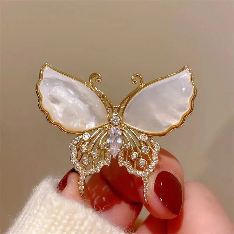 Butterfly-shaped brooch with gold and white design held by a hand with red nail polish.