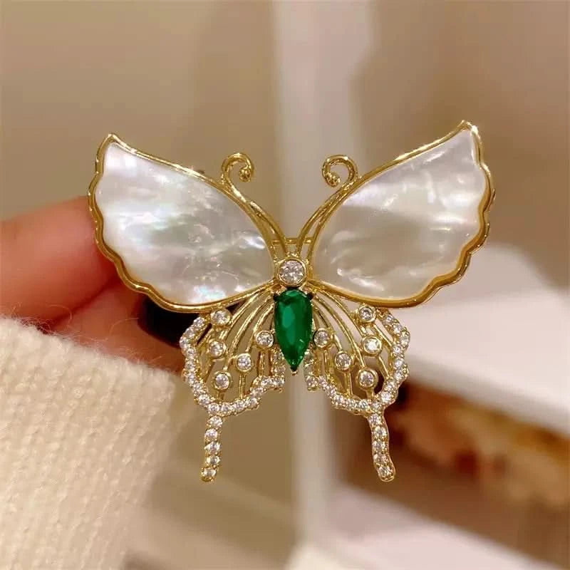 Butterfly-shaped brooch with gold frame, white wings, green center, and diamond-like accents held by a hand.