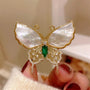 Butterfly-shaped brooch with gold frame, white center, and green gemstone held by a hand.