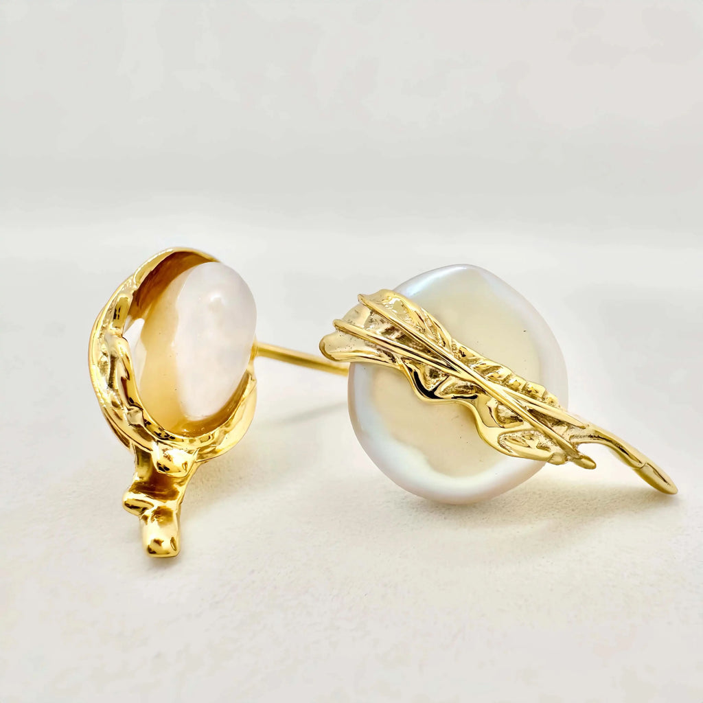 A pair of exquisite earrings featuring large baroque pearls, each embraced by a detailed gold setting with a dynamic, flowing design that evokes a sense of movement. The golden elements wrap around the pearls, highlighting their natural beauty and irregular shapes, set against a light background that enhances the jewelry's luxurious feel.